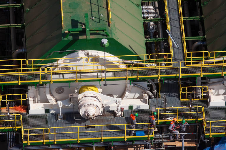 A imagem mostra uma estrutura industrial complexa， com várias plataformas e escadas de metal amarelo e verde. No centro， há um grande equipamento circular com uma parte amarela. Vários trabalhadores， vestidos com uniformes cinzas e coletes de segurança laranja， estão visíveis na plataforma， alguns interagindo entre si. A estrutura parece ser parte de uma instalação de engenharia ou de petróleo.