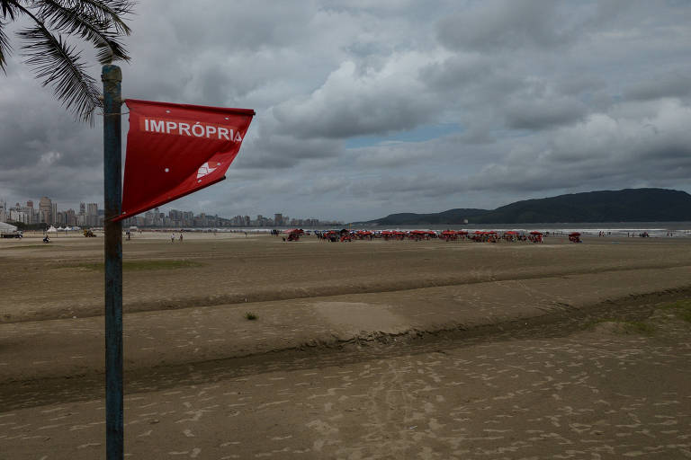 A imagem mostra uma praia com uma bandeira vermelha hasteada， indicando que a água é imprópria para banho. Ao fundo， há uma cidade com prédios e um céu nublado. Na areia， algumas pessoas e guarda-sóis vermelhos são visíveis.