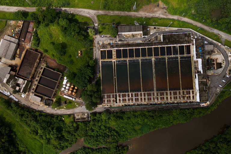 A imagem mostra uma vista aérea de uma estação de tratamento de água. À esquerda, há um edifício de estrutura industrial com telhado escuro e várias áreas de armazenamento. À direita, uma grande estrutura retangular com tanques de água em diferentes tonalidades, cercada por vegetação. Abaixo, um rio flui ao lado da instalação, e a área ao redor é predominantemente verde.