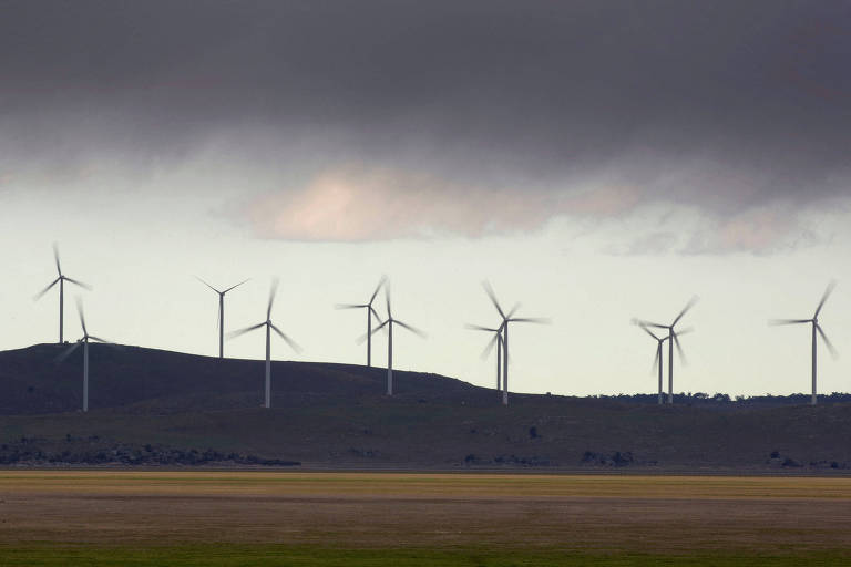 A imagem mostra um parque eólico com várias turbinas eólicas alinhadas em uma colina. O céu está nublado， com nuvens escuras， e a paisagem ao fundo é composta por campos abertos. As turbinas estão em diferentes posições， algumas com as lâminas girando.