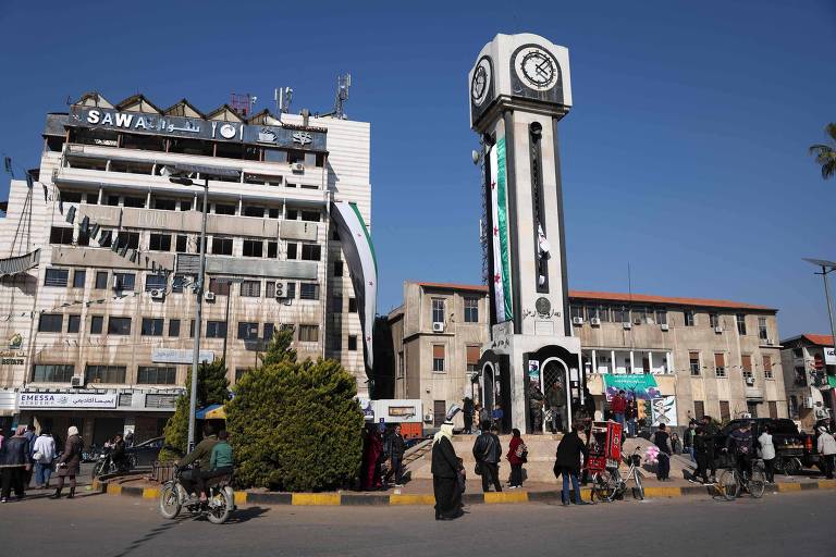 A imagem mostra uma praça com uma torre do relógio central. À esquerda， há um edifício de vários andares com o nome 039;SAWAL039; visível na parte superior. O ambiente é movimentado， com várias pessoas caminhando e algumas em bicicletas. À direita， há um edifício de estilo arquitetônico diferente， com banners e cartazes visíveis. O céu está limpo e azul.