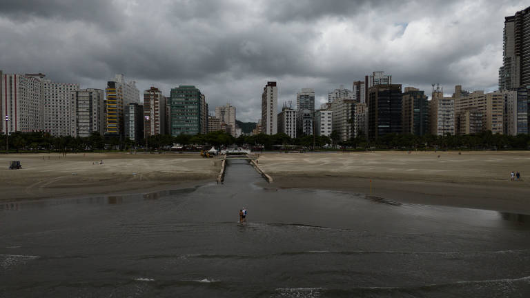 A imagem mostra uma vista panorâmica de uma área urbana com prédios altos e modernos ao fundo， sob um céu nublado e escuro. No primeiro plano， há uma área de areia molhada， de uma praia， refletindo a luz do céu. A atmosfera é de um dia nublado， com nuvens densas cobrindo o céu.