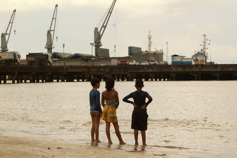 Três crianças em uma praia com píer ao fundo， com guindastes