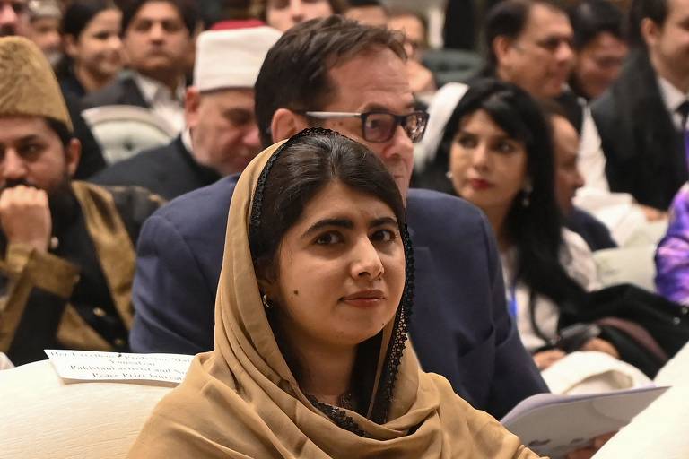 Malala Yousafzai aparece usando um vestido com capuz de cor marrom à frente de outras pessoas que participam de um evento