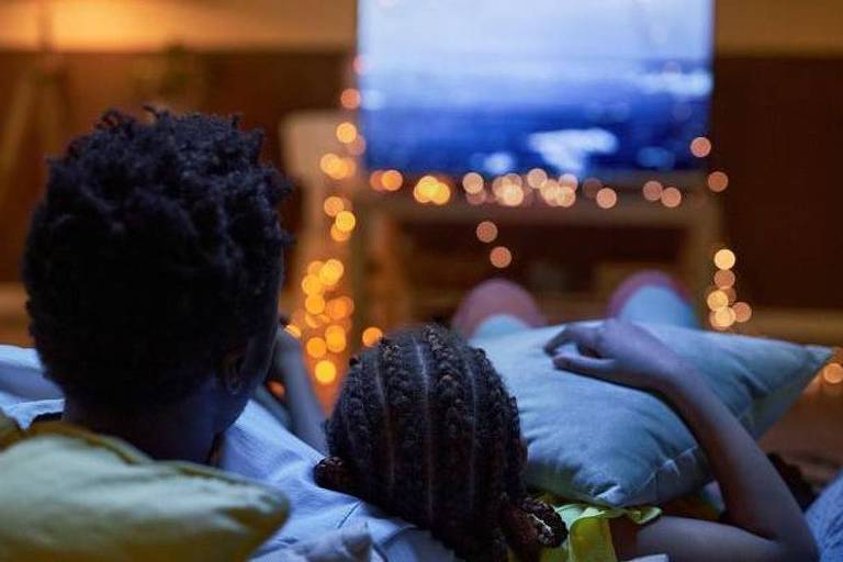 Foto mostra um homem negro com cabelo preto e uma menina negra com tranças pretas， deitados assistindo televisão.