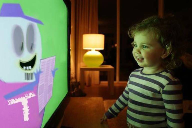 Foto mostra uma menina branca e loira encarando e sorrindo para um personagem animado em uma televisão， com o fundo verde. 