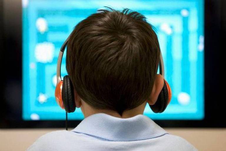 Foto mostra um menino branco de cabelo preto， com fones de ouvido， encarando um computador. O menino está de costas. 