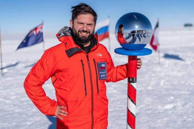 Foto mostra um homem branco de cabelos pretos lisos e barba da mesma cor， sorrindo. Ele veste um agasalho laranja e segura um mastro， com um globo de vidro espelhado na ponta， com a mão esquerda. Ele está em um ambiente nevado. 