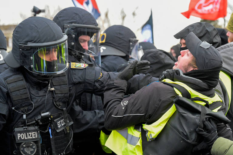 A imagem mostra um confronto entre policiais e manifestantes. Os policiais estão vestidos com uniformes pretos e capacetes， alguns com visores. Eles estão em posição defensiva， enquanto um manifestante， vestido com um colete amarelo， parece estar em uma situação de tensão， gesticulando e se aproximando dos policiais. Ao fundo， há bandeiras e outros manifestantes.