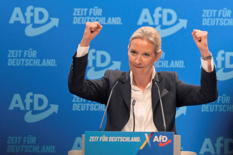 Uma mulher em pé， com os braços levantados em sinal de celebração， fala em um evento político. Ela está vestida com um blazer escuro e uma blusa clara. Ao fundo， há um painel azul com o logotipo de um partido político e uma frase que diz， em alemão， 039;A hora da Alemanha039;.