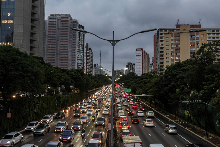 A imagem mostra uma vista de uma avenida movimentada em uma cidade， com muitos carros em movimento. O céu está nublado， indicando que pode chover. À esquerda， há prédios altos e à direita， edifícios residenciais. Árvores estão visíveis ao longo da avenida， e as luzes dos carros estão acesas devido à falta de luz natural.