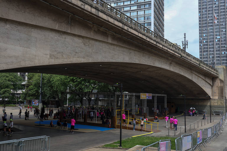 A imagem mostra um espaço amplo sob um viaduto, com várias pessoas reunidas. Há uma área de recreação com brinquedos e um piso azul. Algumas pessoas estão vestidas com camisetas rosas. Ao fundo, há prédios altos e árvores ao redor.