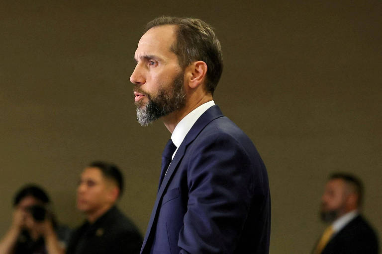 Um homem com barba e cabelo curto， vestido com um terno escuro e gravata， está em perfil. Ele parece estar em um ambiente forma. Ao fundo， há algumas pessoas， mas não estão claramente visíveis