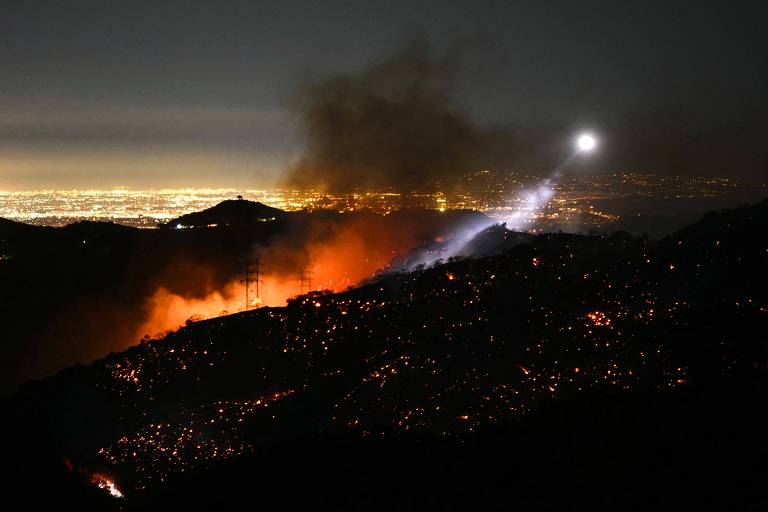 A imagem mostra um incêndio florestal em uma montanha durante a noite. Chamas laranjas e fumaça escura se elevam， enquanto ao fundo é possível ver a iluminação da cidade. Uma luz branca， de um helicóptero， ilumina a cena.