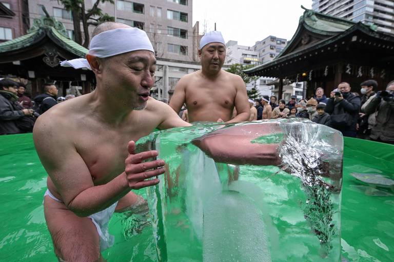 Dois homens sem camisa, usando faixas brancas na cabeça, estão em frente a um bloco de gelo em um ambiente ao ar livre. Eles parecem estar participando de uma cerimônia ou evento tradicional, com um templo ao fundo e uma multidão de espectadores observando. O cenário é urbano, com prédios visíveis ao fundo.