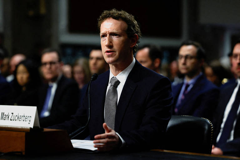 Imagem mostra Mark Zuckerberg， um homem de terno escuro e gravata clara que está sentado à mesa em uma audiência no Senado. Ele parece estar falando ou respondendo a perguntas， com uma expressão séria. Ao fundo， há várias pessoas sentadas， algumas olhando atentamente. Na mesa à sua frente， há um cartão com o seu nome.