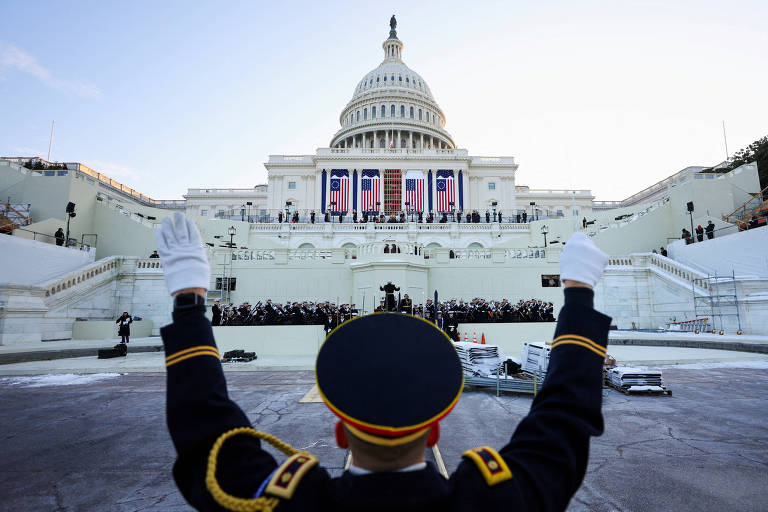 A imagem mostra um oficial em uniforme militar， de costas， levantando as mãos em direção ao Capitólio dos Estados Unidos. O edifício está ao fundo， decorado com bandeiras e uma multidão de pessoas visíveis nas escadas. O céu está claro， indicando um dia ensolarado.