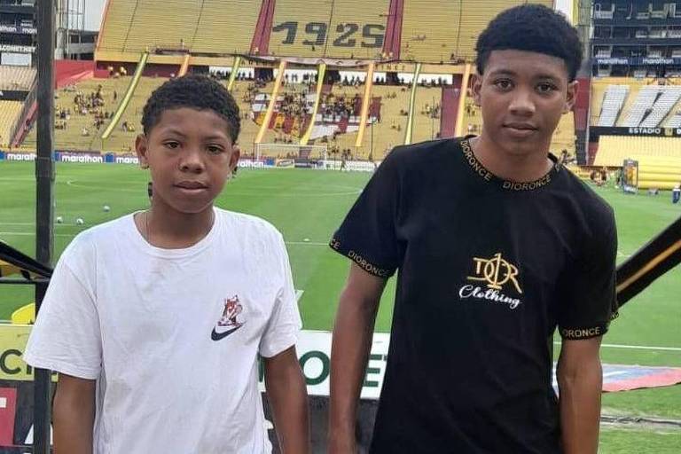 Foto mostra dois garotos negros pousando para o retrato dentro de um estádio de futebol. O da direita usa uma camiseta preta e é mais alto que o da esquerda， que usa uma camiseta branca. 