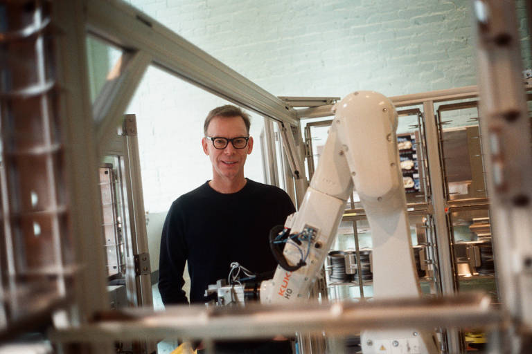 A imagem mostra um homem usando óculos e uma camiseta preta， posicionado em um ambiente industrial com um braço robótico ao seu lado. O robô está em uma estrutura metálica， e o fundo é composto por equipamentos e maquinário.