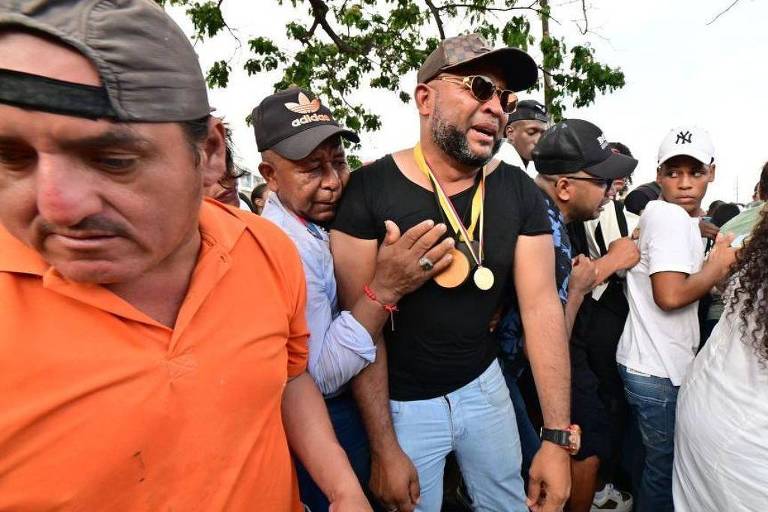 Foto mostra um homem negro de camiseta preta， boné， óculos escuros e várias medalhas no peito. Ele chora e é consolado por uma multidão de pessoas da mesma cor dele. 