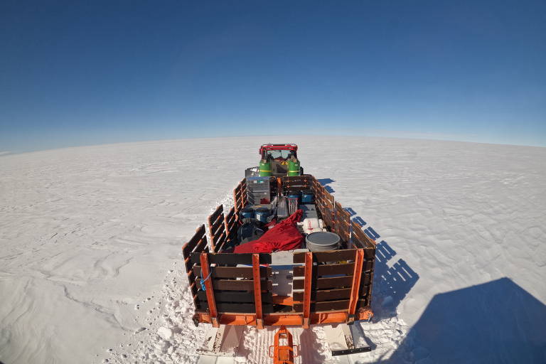 A imagem mostra uma vista de trás de um veículo de transporte em uma vasta área coberta de neve. O veículo está puxando uma plataforma com diversos itens, incluindo barris, uma caixa verde e um cobertor vermelho. O céu é claro e azul, e a superfície da neve se estende até o horizonte, sem obstáculos visíveis.