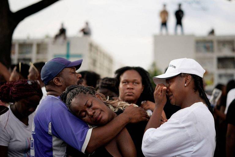 Foto mostra três mulheres e um homem， todos negros， chorando num velório. ao redor deles， há outras pessoas 