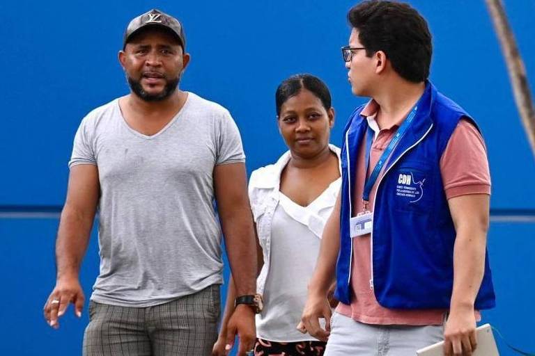 Foto mostra um homem negro de camiseta cinza， com barba preta e um boné， também preto. Ao lado dele， uma mulher negra de cabelo liso preto e camiseta branca. A dupla é acompanhada de um homem branco， com camisa polo vermelha e um colete azul， com um chachá. Ele tem cabelos pretos lisos e usa óculos. 