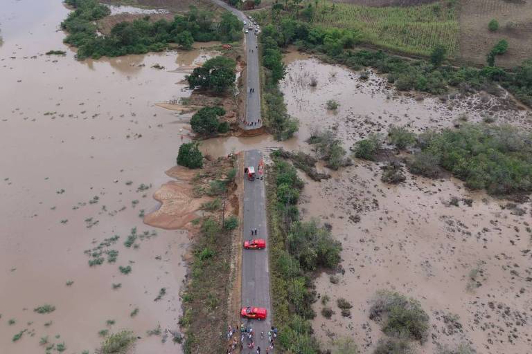 A imagem mostra uma estrada que se estende por uma área alagada， com água em ambos os lados. O cenário inclui campos verdes e uma paisagem montanhosa ao fundo. Há alguns veículos visíveis na estrada， e a água parece ter invadido as margens， criando uma grande extensão de alagamento.