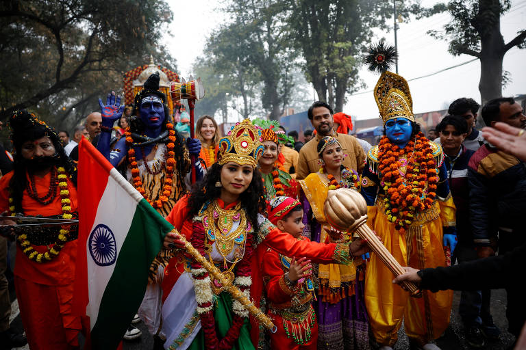 A imagem mostra um grupo de pessoas participando de uma celebração cultural na Índia. Algumas pessoas estão vestidas com trajes tradicionais coloridos, incluindo figuras que representam deuses hindus, como Krishna e outros. Uma mulher na frente segura a bandeira da Índia, enquanto uma criança segura um objeto em forma de bastão. O ambiente é festivo, com árvores ao fundo e uma multidão de espectadores.