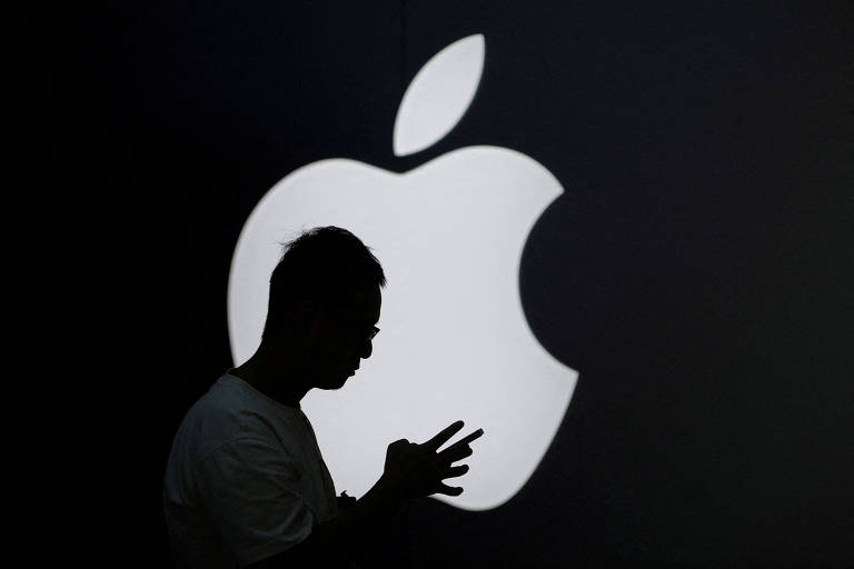 A imagem mostra a silhueta de uma pessoa em frente a um grande logotipo da Apple， que é iluminado em branco contra um fundo escuro. A pessoa parece estar usando um dispositivo móvel， com as mãos posicionadas em frente ao corpo.