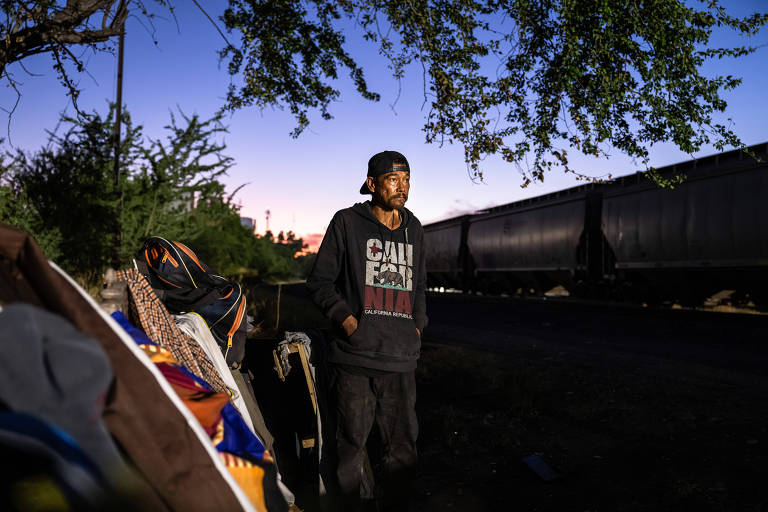  A imagem mostra um homem de pé perto de trilhos de trem, em um ambiente ao ar livre. Ele usa um moletom com o logotipo da Califórnia e está em uma posição relaxada, com as mãos nos bolsos. O ambiente parece ser uma área de espera, com mochilas e cobertores ao fundo, à medida que o sol se põe.