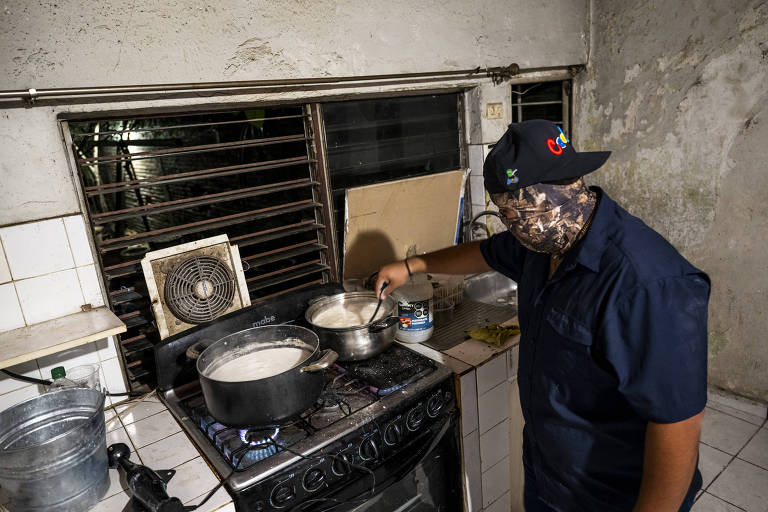 Um homem está em uma cozinha simples, mexendo algo em uma panela sobre o fogão. Ele usa uma máscara camuflada e um boné, enquanto cozinha o que parece ser uma substância em dois potes. O ambiente está pouco iluminado, com um ventilador de parede visível e janelas com barras.