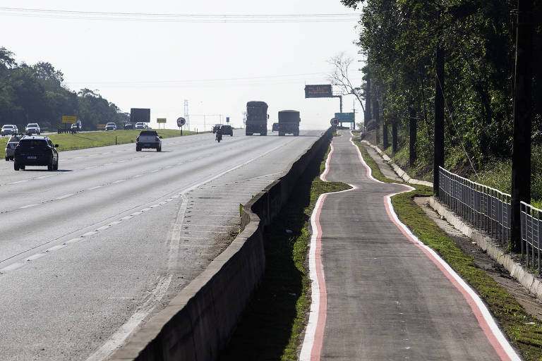 A imagem mostra uma rodovia com várias faixas de tráfego， onde veículos estão se deslocando. À direita， há uma calçada ou ciclovia， que é mais estreita e possui uma borda pintada de vermelho. A vegetação está presente ao longo da calçada， e o céu está claro ao fundo.