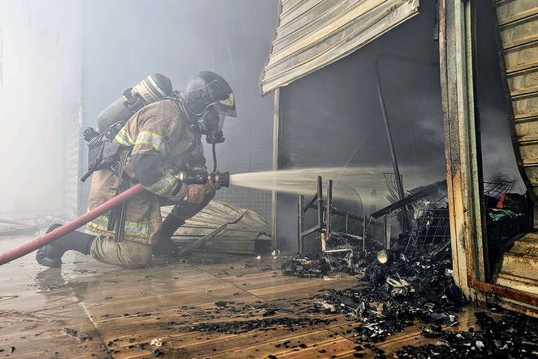 Um bombeiro está agachado， usando um equipamento de proteção， enquanto combate um incêndio em um ambiente com fumaça densa. Ele direciona um jato de água para uma área que parece ter sido queimada， com restos carbonizados visíveis no chão. A estrutura ao fundo é uma parede de madeira com tábuas expostas.