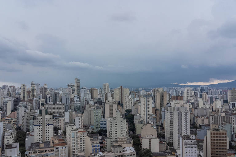 A imagem mostra uma vista panorâmica de uma cidade com muitos edifícios altos. O céu está nublado， com tons de cinza， e há uma leve iluminação ao longe， sugerindo que o sol está se pondo. A cidade parece densa， com uma variedade de estruturas arquitetônicas visíveis.