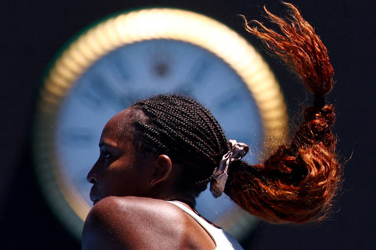 A imagem mostra uma jogadora de tênis de perfil, com cabelo trançado e uma parte do cabelo solto, que se destaca em um fundo desfocado. Ao fundo, há um relógio grande com um design circular e detalhes em verde e dourado.