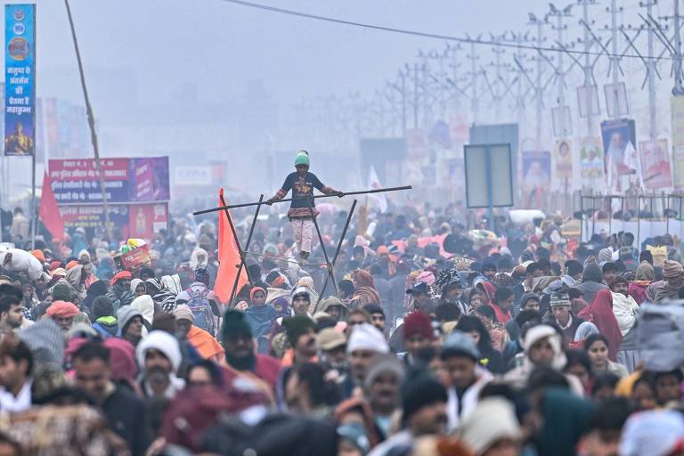 A imagem mostra uma grande multidão reunida em um evento ao ar livre, com pessoas vestindo roupas variadas, algumas com cobertores e chapéus devido ao frio. No centro, um jovem equilibrista está em cima de uma corda, segurando varas para se equilibrar. Ao fundo, há várias placas e banners, e a atmosfera parece enevoada, sugerindo um clima frio e possivelmente festivo.