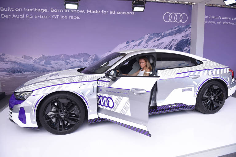 A imagem mostra um carro da marca Audi， modelo elétrico， estacionado em um evento. O veículo é predominantemente branco com detalhes em roxo e possui uma porta aberta. Dentro do carro， há uma pessoa visível. Ao fundo， há uma parede com o logotipo da Audi e texto em português que menciona a marca e o modelo do carro.