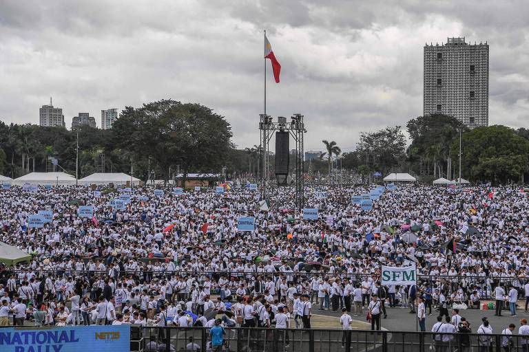 A imagem mostra uma grande multidão reunida em uma área pública， com pessoas vestindo camisetas brancas. No centro， há um mastro com a bandeira das Filipinas. Ao fundo， arranha-céus e árvores são visíveis， e tendas brancas estão montadas ao longo da área. Algumas pessoas seguram cartazes e bandeiras coloridas， enquanto outras estão em pé ou sentadas. O céu está nublado.