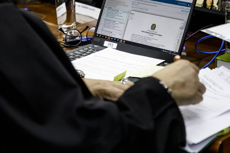 A imagem mostra uma pessoa com uma roupa escura， possivelmente um manto， usando um computador portátil. A mão da pessoa está segurando um objeto enquanto escreve em papéis sobre uma mesa. O computador exibe uma tela com informações visíveis， e há outros itens de escritório ao redor， como canetas e documentos.