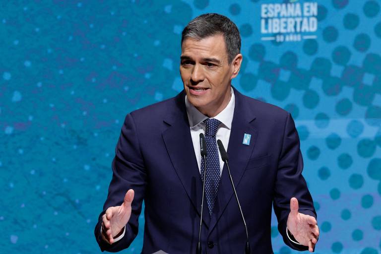 A imagem mostra Pedro Sanchez， um homem em um terno escuro que está falando em um evento， gesticulando com as mãos. O fundo é azul com padrões circulares e contém o texto 039;ESPAÑA EN LIBERTAD039;.