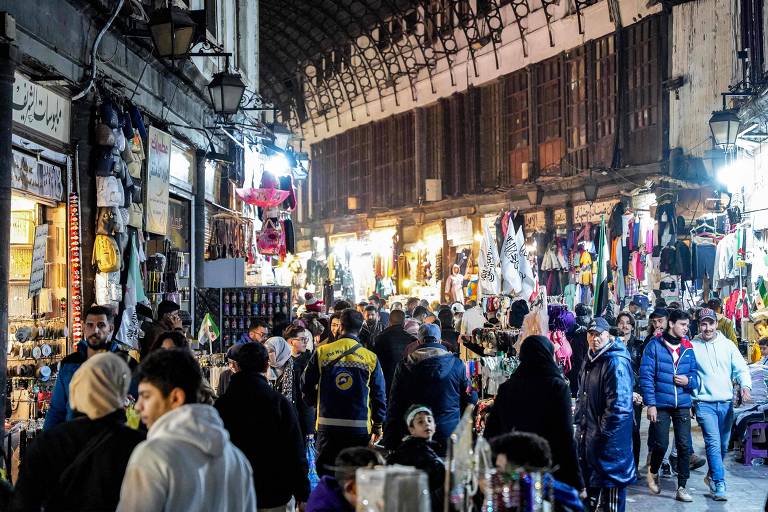 A imagem mostra um mercado noturno em Damasco， com várias barracas e lojas iluminadas. Há muitas pessoas caminhando pelas ruas， algumas paradas para olhar produtos. As lojas vendem roupas， acessórios e outros itens. O ambiente é movimentado e vibrante， com uma arquitetura tradicional visível ao fundo.
