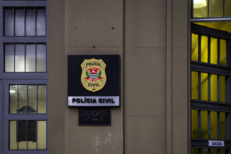 A imagem mostra uma placa da Polícia Civil fixada em uma parede. A placa possui um escudo com cores verde， vermelho e amarelo， e o texto 039;POLÍCIA CIVIL039; em letras grandes e em negrito. Abaixo， há um número 039;527039;. Ao fundo， é possível ver janelas com grades e uma porta com a palavra 039;SAÍDA039; visível.