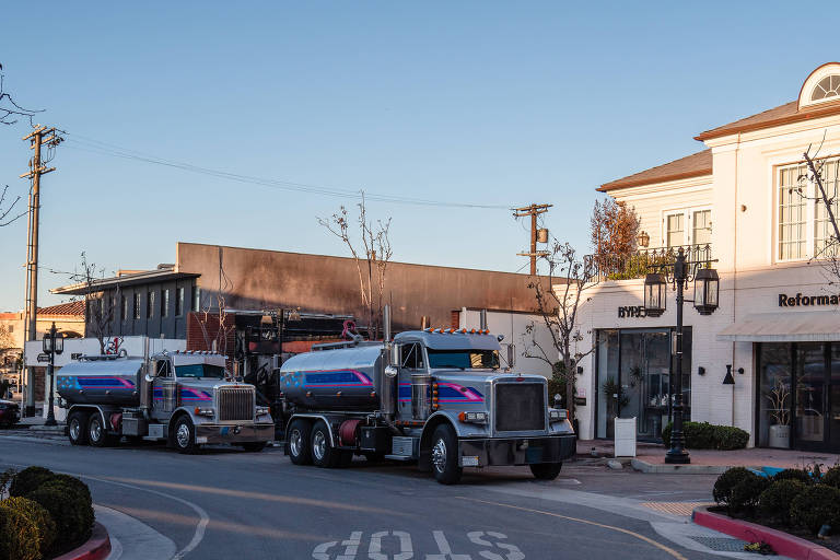 A imagem mostra dois caminhões-tanque estacionados em uma rua comercial. O primeiro caminhão， à esquerda， é prateado com detalhes em azul e rosa. Ao fundo， há um edifício com uma fachada escura， possivelmente danificada， e um outro edifício branco com janelas e uma placa que diz 039;Reformation039;. A rua é pavimentada e há uma sinalização de 039;STOP039; visível no chão. Árvores sem folhas estão presentes ao longo da calçada.