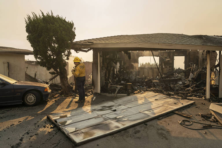 A imagem mostra um bombeiro em uma área devastada por incêndio， ao lado de um carro azul e uma árvore. O fundo revela uma estrutura de casa que foi severamente danificada， com paredes queimadas e escombros visíveis. O céu está nublado， indicando fumaça ou poluição， e há uma porta de garagem caída no chão.