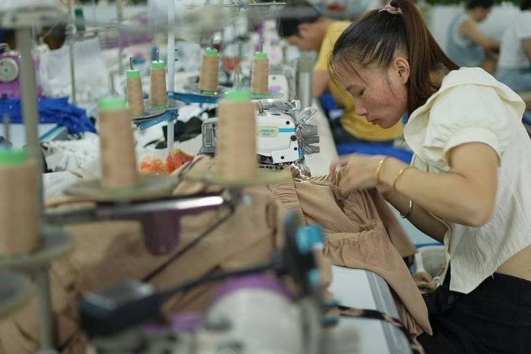 A imagem mostra uma mulher trabalhando em uma linha de produção de uma fábrica de confecção. Ela está concentrada em costurar um tecido， com várias máquinas de costura e carretéis de linha ao fundo. O ambiente é bem iluminado e organizado， com outros trabalhadores visíveis em segundo plano.