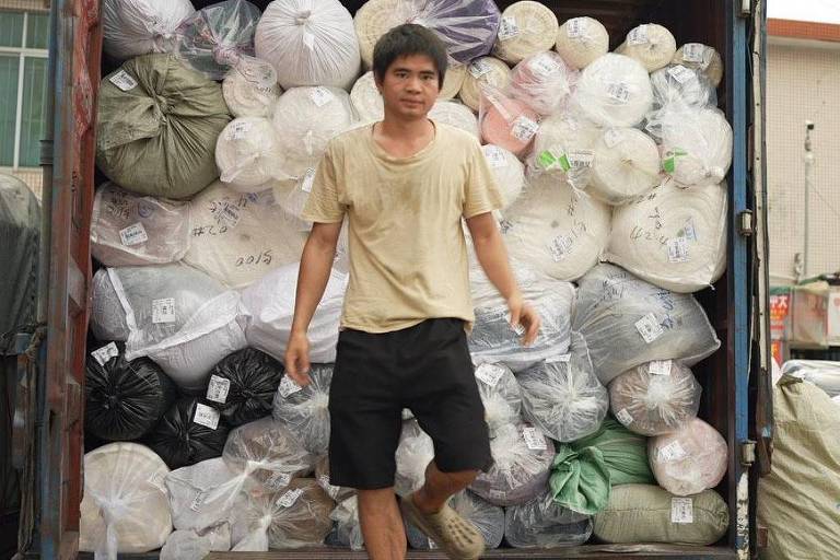 Um homem de camiseta bege e shorts preto está descendo de um caminhão empilhado com rolos de tecido de várias cores: rosa， branco， verde.
