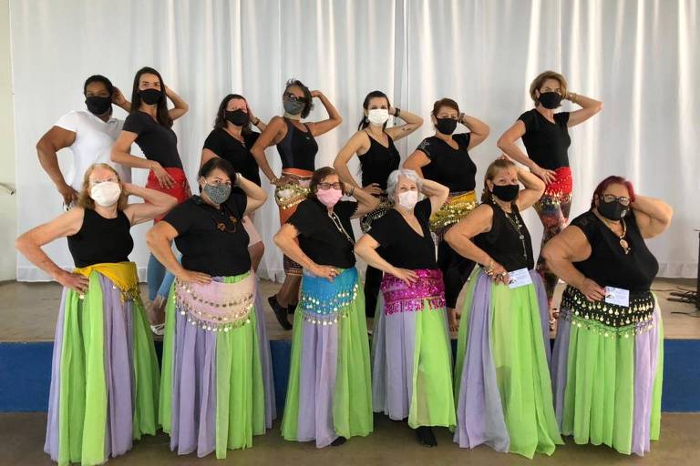 Um grupo de mulheres está posando para a foto em um palco. Elas estão usando camisetas pretas e saias longas de cores variadas， como verde， lilás e rosa. Todas estão usando máscaras faciais. O fundo é uma cortina branca