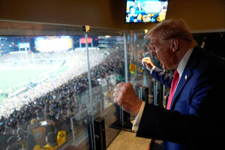 A imagem mostra um homem em um palanque assistindo a um evento esportivo. Ele está vestido com um terno azul e uma gravata vermelha， gesticulando com as mãos em um momento de entusiasmo. Ao fundo， há uma grande multidão assistindo ao jogo， com luzes e telas visíveis. O ambiente parece ser uma área VIP， com uma vista panorâmica do campo.