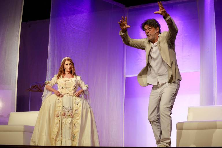 A imagem mostra uma cena de teatro com dois atores em um palco. À esquerda, uma mulher com cabelo castanho claro e longo, vestindo um vestido amarelo com detalhes florais, está em uma pose de descontentamento, com as mãos na cintura. À direita, um homem com cabelo grisalho e barba, vestido com um terno claro, está gesticulando de forma expressiva. O fundo do palco é iluminado em tons de roxo, e há uma estrutura de pano que divide o espaço.